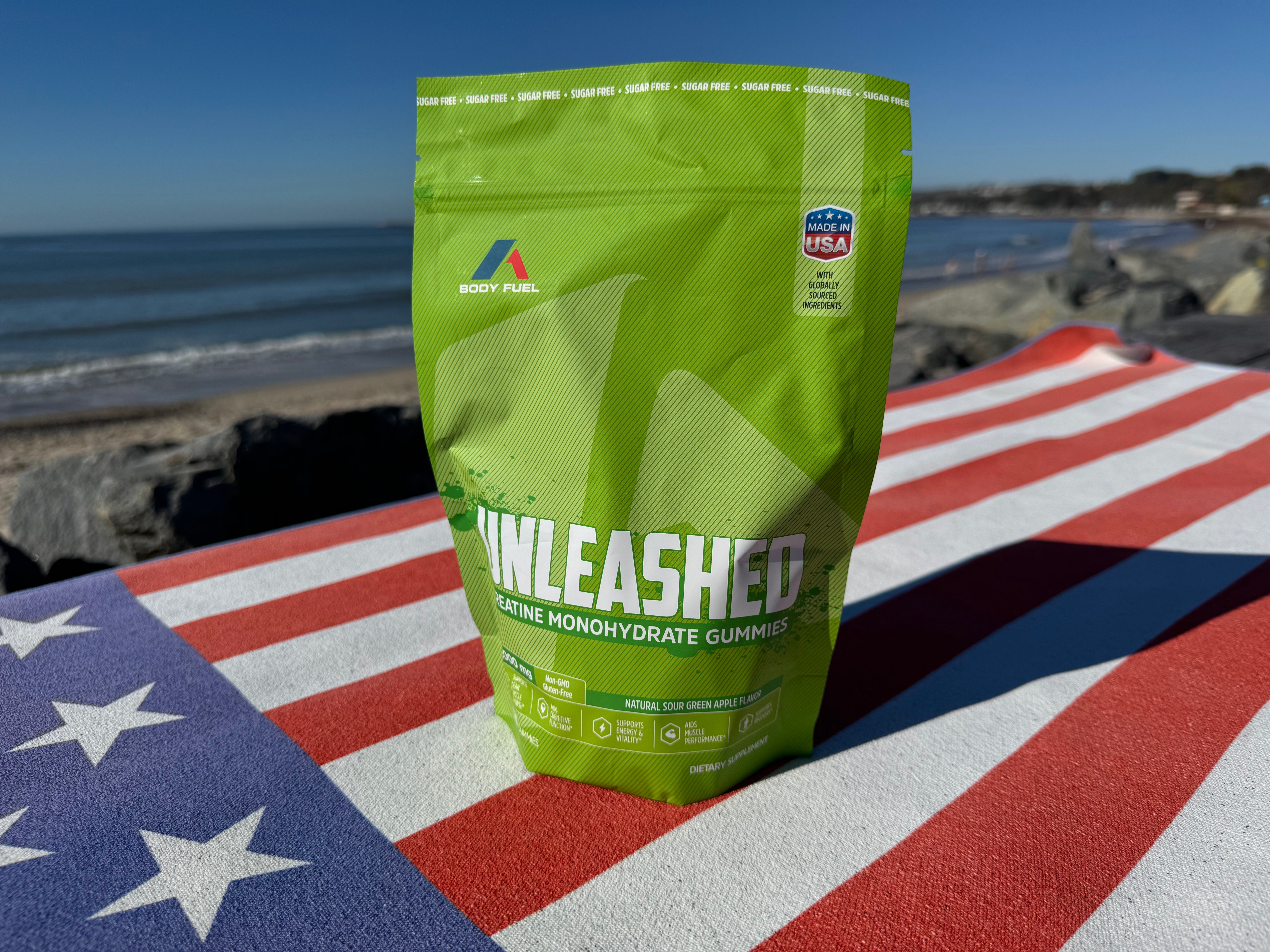 Green 'Unleashed Creatine Gummies Sugar Free-Green Apple' supplement package on a U.S. flag with a beach background